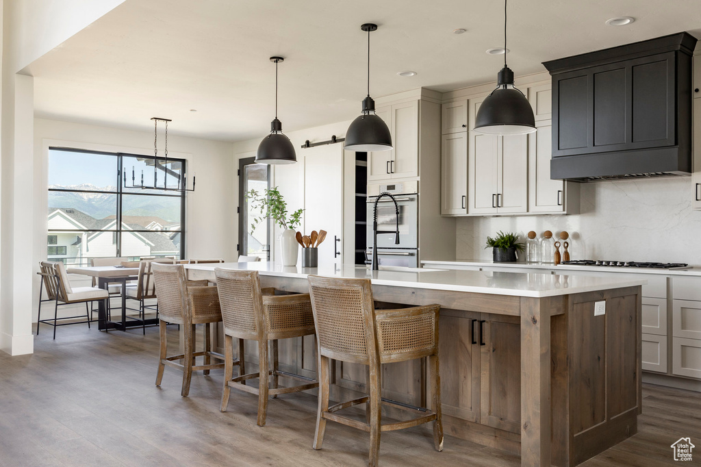 Kitchen featuring wood finished floors, an island with sink, backsplash, light countertops, and gas stovetop