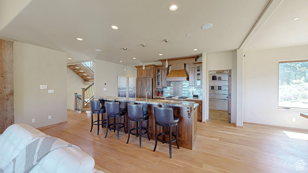 Kitchen featuring custom exhaust hood, light wood-style floors, an island with sink, a breakfast bar, and tasteful backsplash