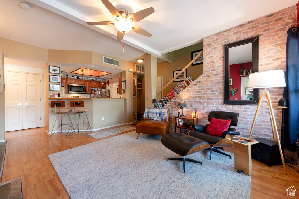 Living room featuring wood finished floors, a ceiling fan, brick wall, baseboards, and stairs