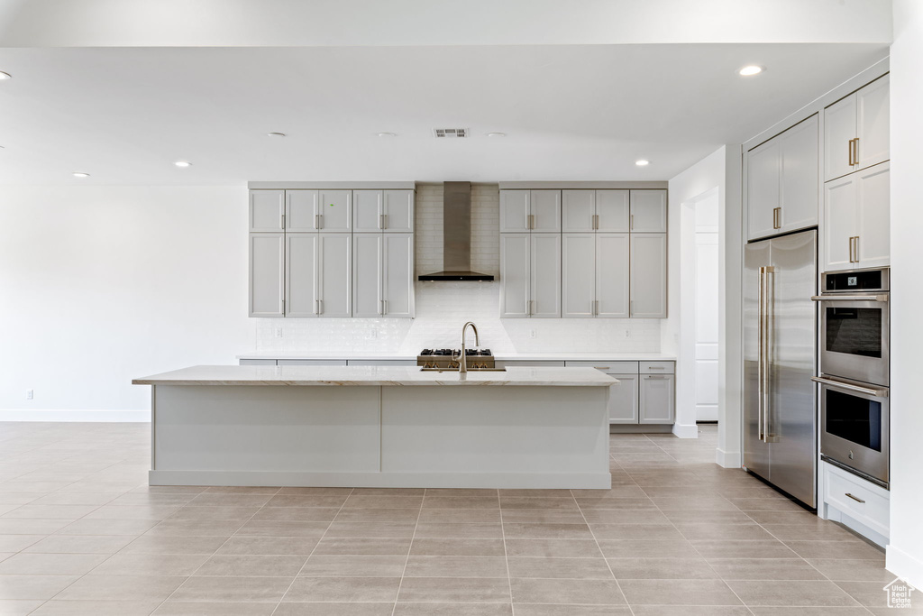 Kitchen featuring wall chimney exhaust hood, stainless steel appliances, an island with sink, recessed lighting, and light tile patterned floors