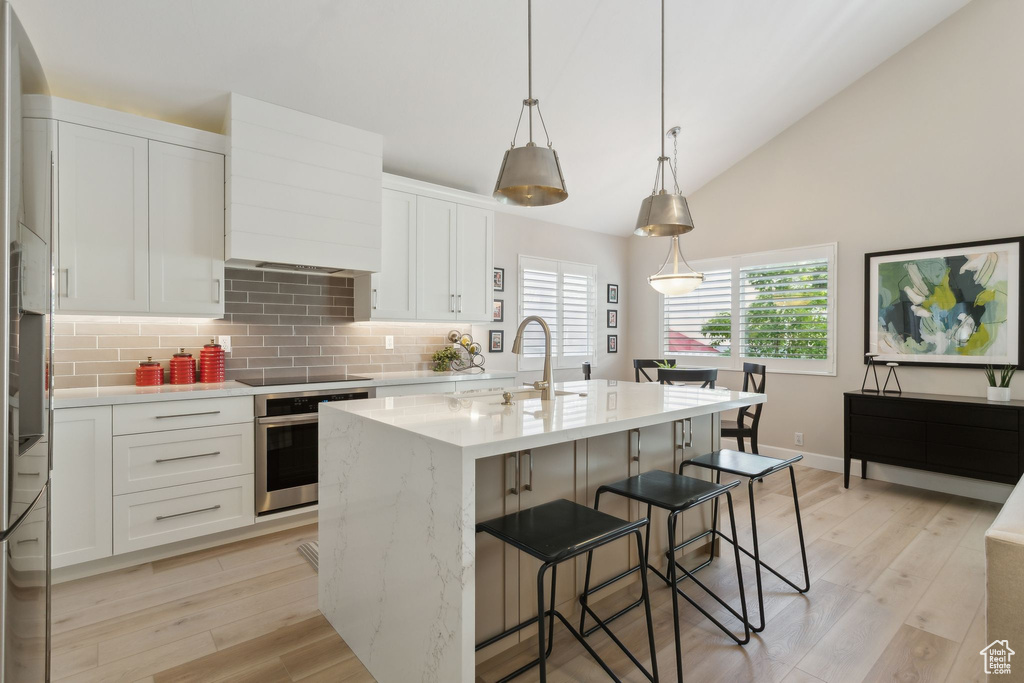 Kitchen featuring stainless steel oven, backsplash, black electric stovetop, a center island with sink, and light wood-style flooring