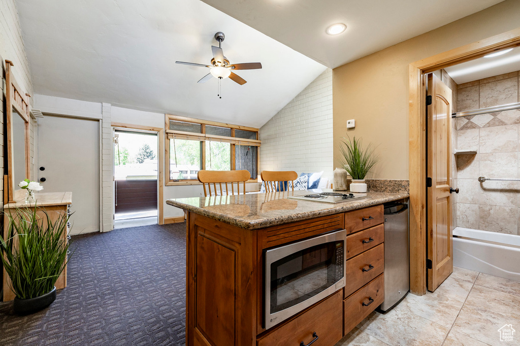 Kitchen with stainless steel microwave, brown cabinetry, a ceiling fan, vaulted ceiling, and light stone countertops