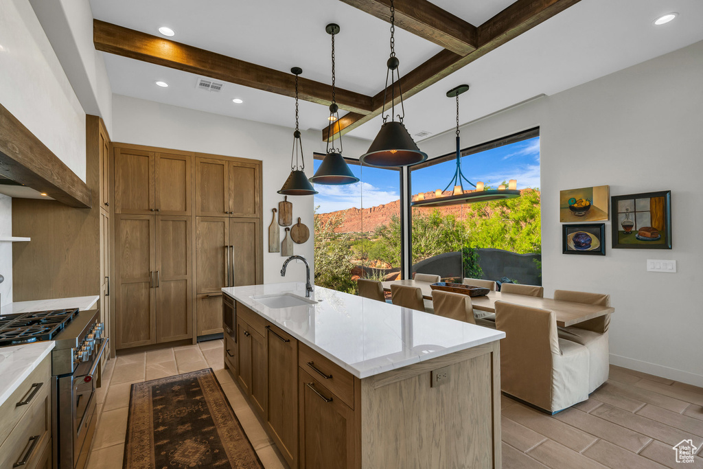 Kitchen with a sink, high end stove, beam ceiling, brown cabinetry, and decorative light fixtures