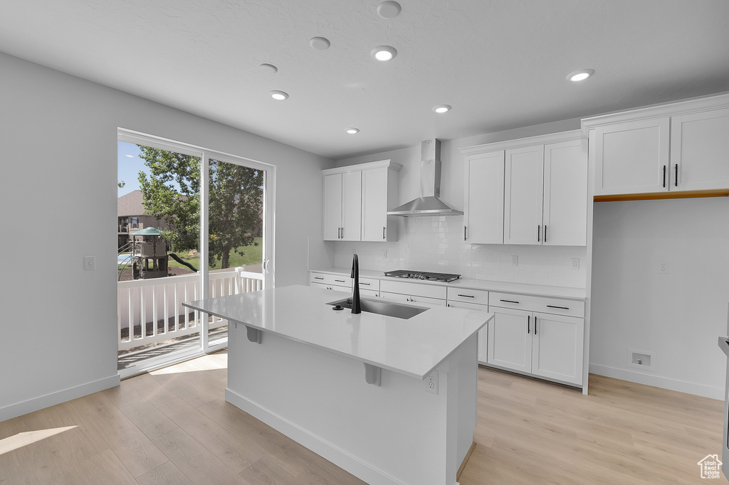 Kitchen featuring wall chimney exhaust hood, a sink, light wood-style floors, a center island with sink, and recessed lighting