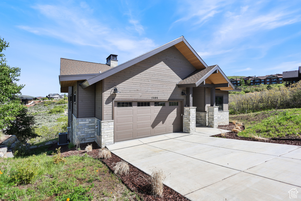 View of front of property featuring stone siding, concrete driveway, an attached garage, a chimney, and roof with shingles