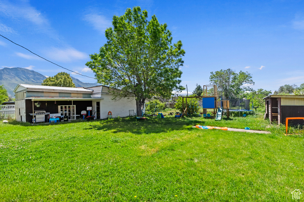 View of grassy yard featuring a trampoline, a playground, a mountain view, and an outbuilding