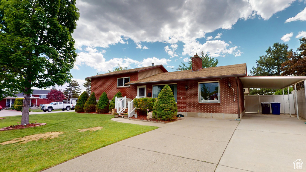 Split level home featuring concrete driveway, brick siding, a chimney, an attached carport, and a front lawn