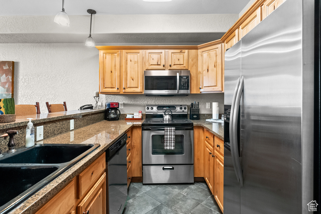 Kitchen with stainless steel appliances, a sink, a textured wall, hanging light fixtures, and dark tile patterned flooring