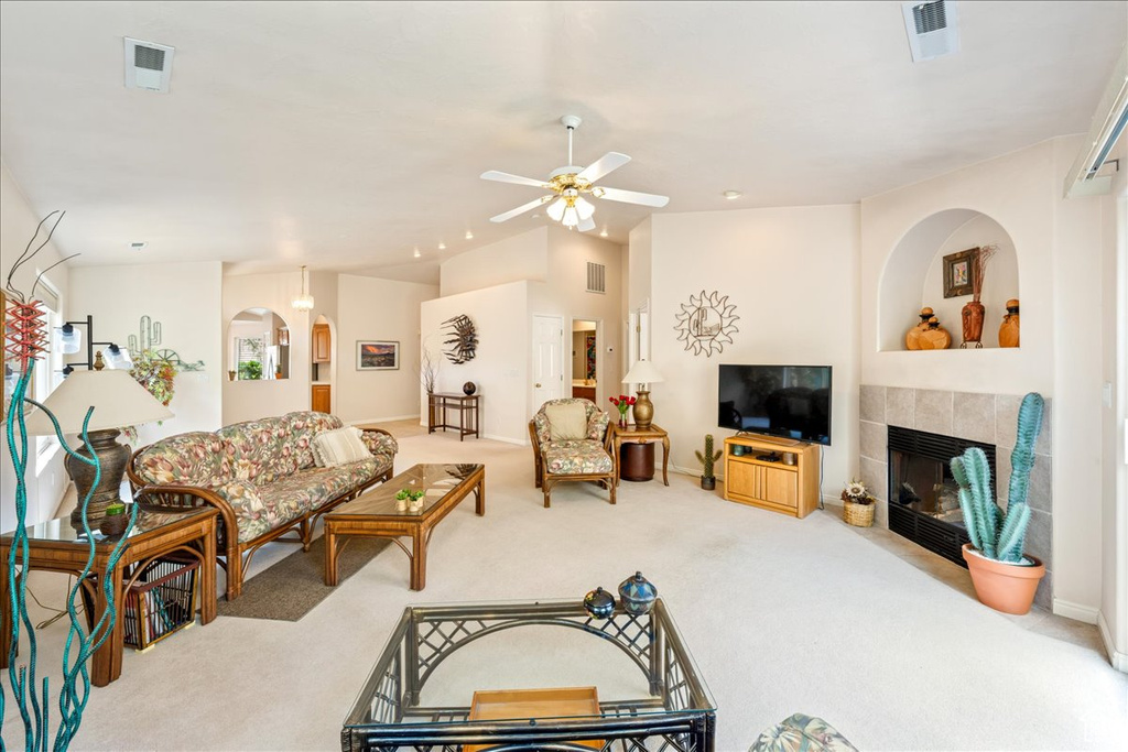 Carpeted living room with ceiling fan, a tiled fireplace, vaulted ceiling, arched walkways, and baseboards