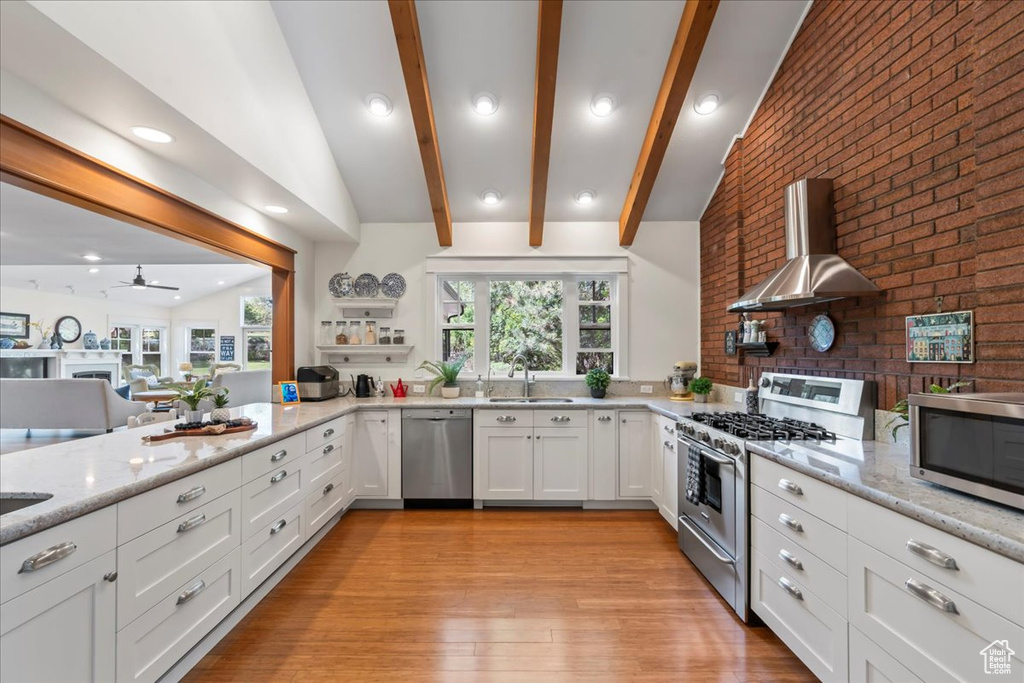 Kitchen with appliances with stainless steel finishes, wall chimney range hood, light stone countertops, a sink, and white cabinets