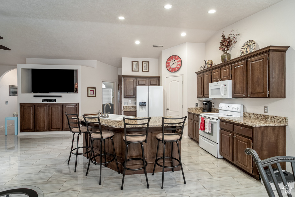 Kitchen with white appliances, a breakfast bar area, a center island with sink, light stone countertops, and recessed lighting