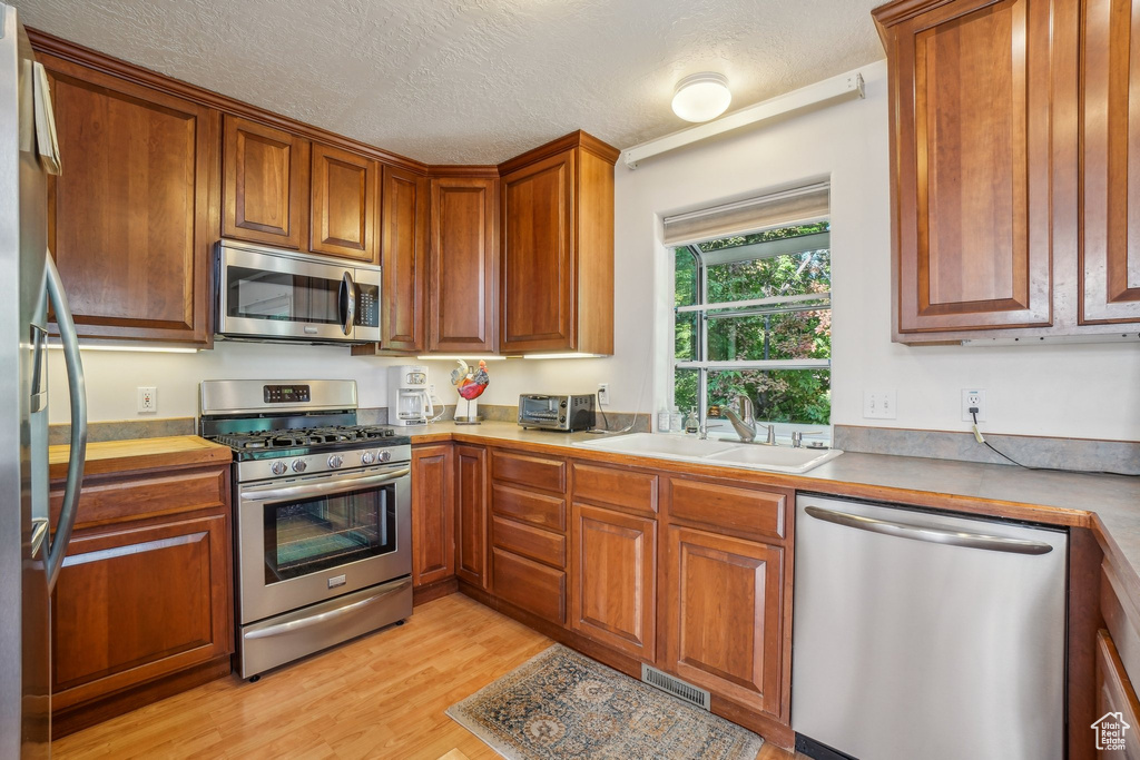 Kitchen with appliances with stainless steel finishes, a sink, light wood-type flooring, brown cabinetry, and a textured ceiling