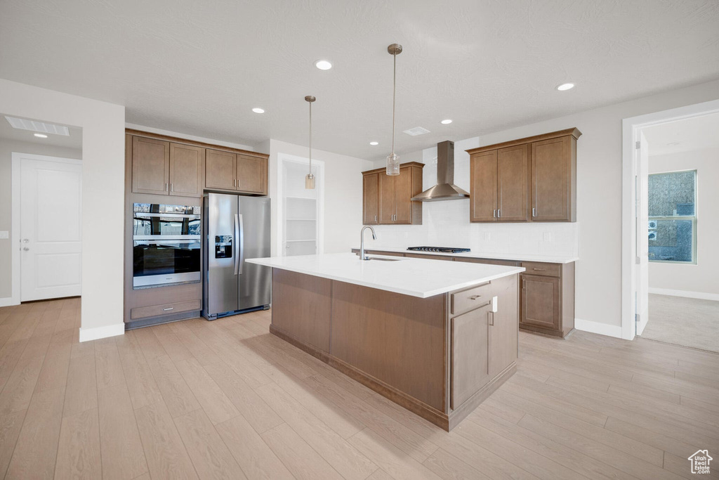 Kitchen with wall chimney exhaust hood, appliances with stainless steel finishes, light wood-type flooring, a center island with sink, and recessed lighting
