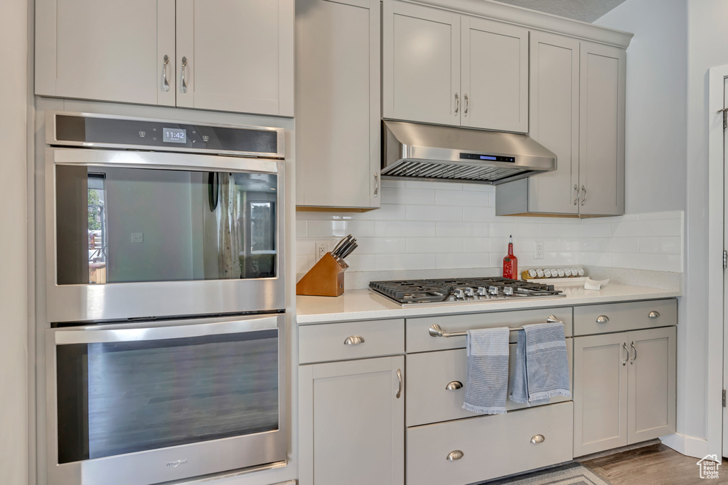 Kitchen with under cabinet range hood, stainless steel appliances, light countertops, tasteful backsplash, and gray cabinetry