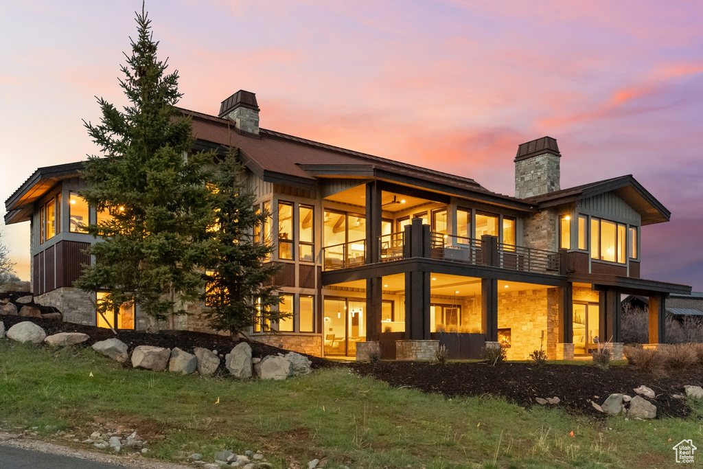 Back of house at dusk with stone siding, a chimney, and a balcony