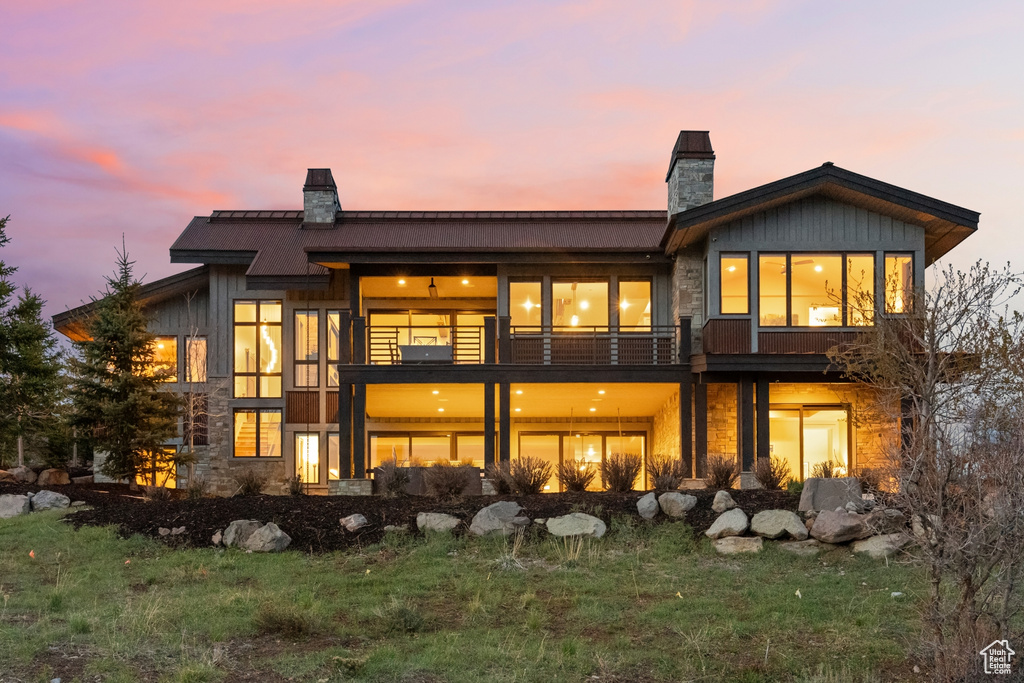 Back of house at dusk with a balcony, a chimney, and stone siding