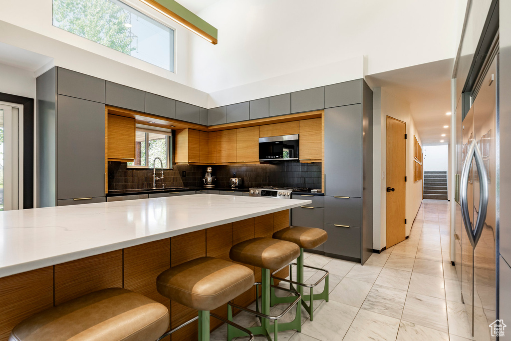 Kitchen featuring appliances with stainless steel finishes, a kitchen breakfast bar, decorative backsplash, gray cabinets, and a towering ceiling