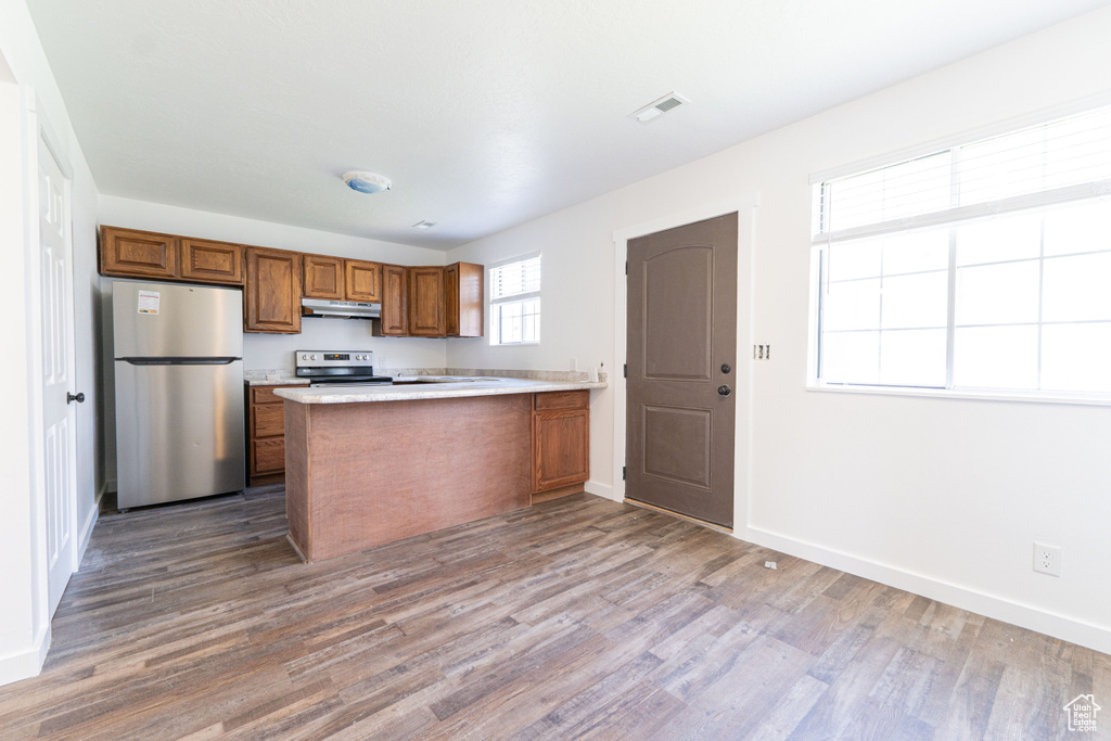 Kitchen with appliances with stainless steel finishes, under cabinet range hood, brown cabinets, wood finished floors, and a peninsula