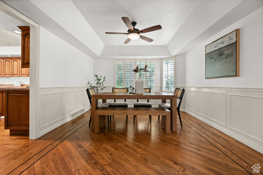 Dining room with ceiling fan, a raised ceiling, light wood finished floors, and a wainscoted wall