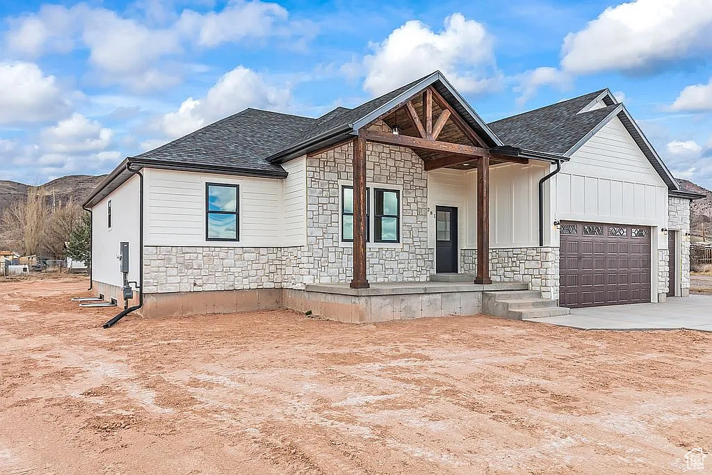 View of front of home with a garage, stone siding, driveway, covered porch, and roof with shingles