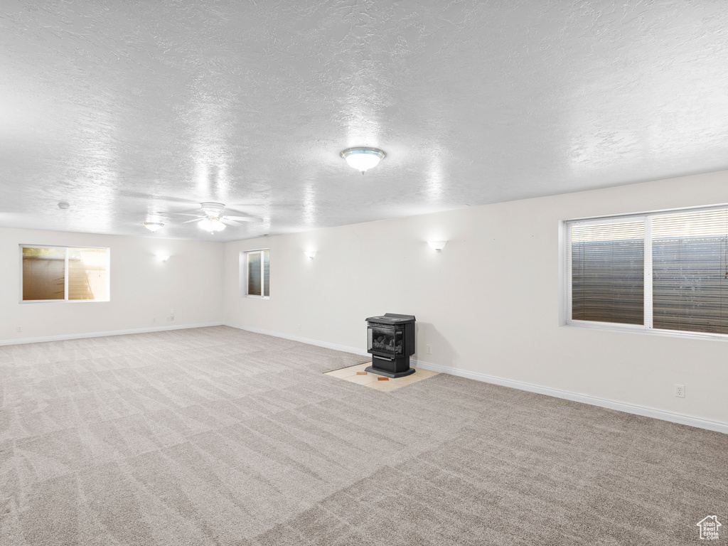 Carpeted empty room with a wood stove, a textured ceiling, a ceiling fan, healthy amount of natural light, and baseboards