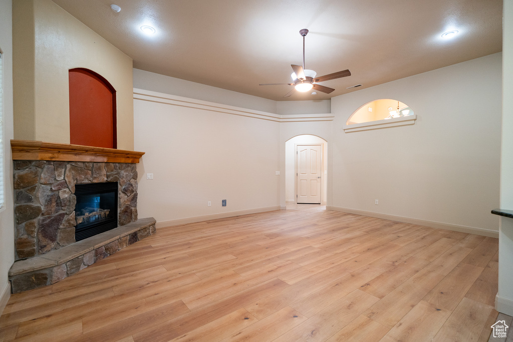 Unfurnished living room featuring a ceiling fan, a stone fireplace, light wood-type flooring, arched walkways, and baseboards