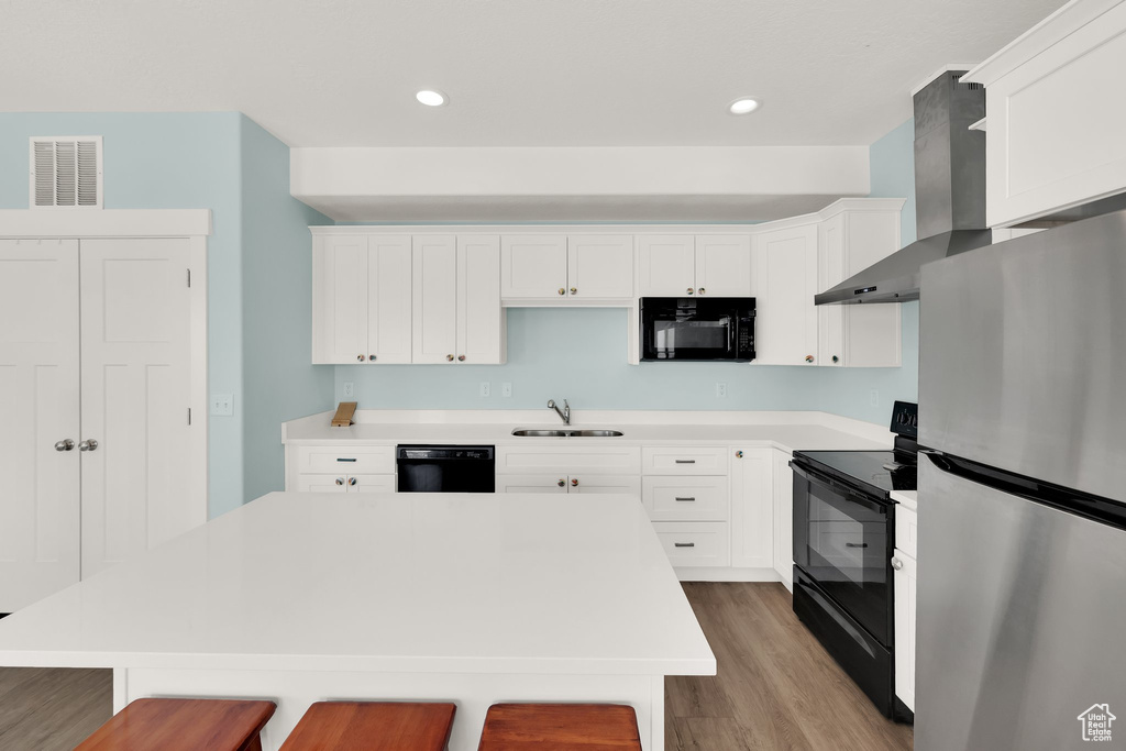 Kitchen with black appliances, wall chimney exhaust hood, light wood-type flooring, a breakfast bar area, and a sink