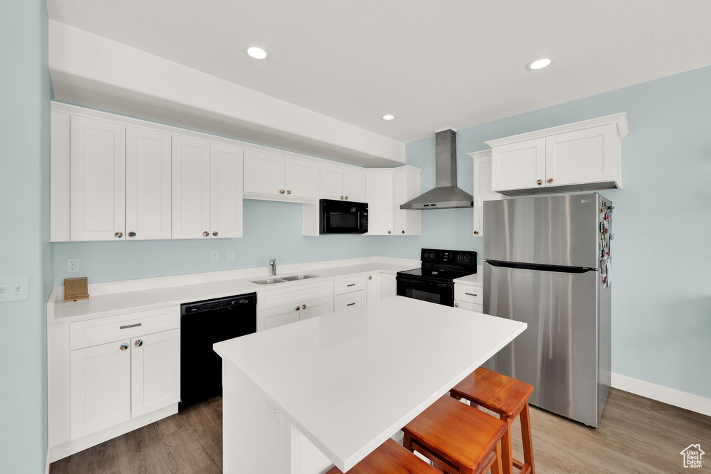 Kitchen with black appliances, wall chimney exhaust hood, a sink, a breakfast bar, and light wood finished floors