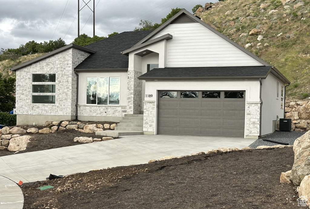 View of front of home featuring stone siding, driveway, cooling unit, and a shingled roof