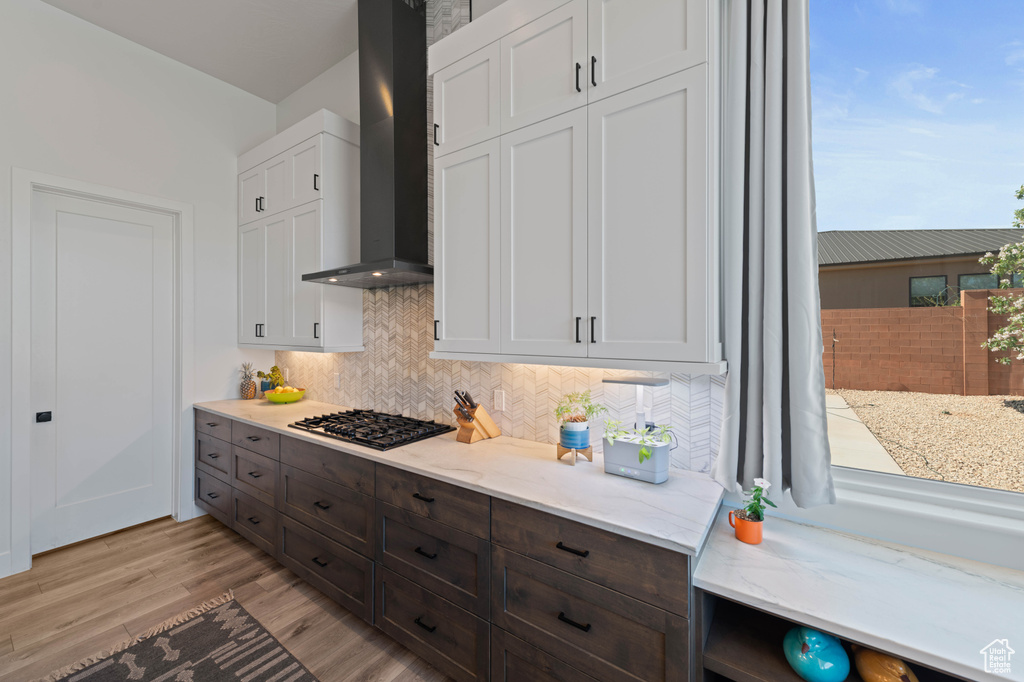 Kitchen with wall chimney exhaust hood, black gas cooktop, decorative backsplash, light wood-style flooring, and light stone countertops