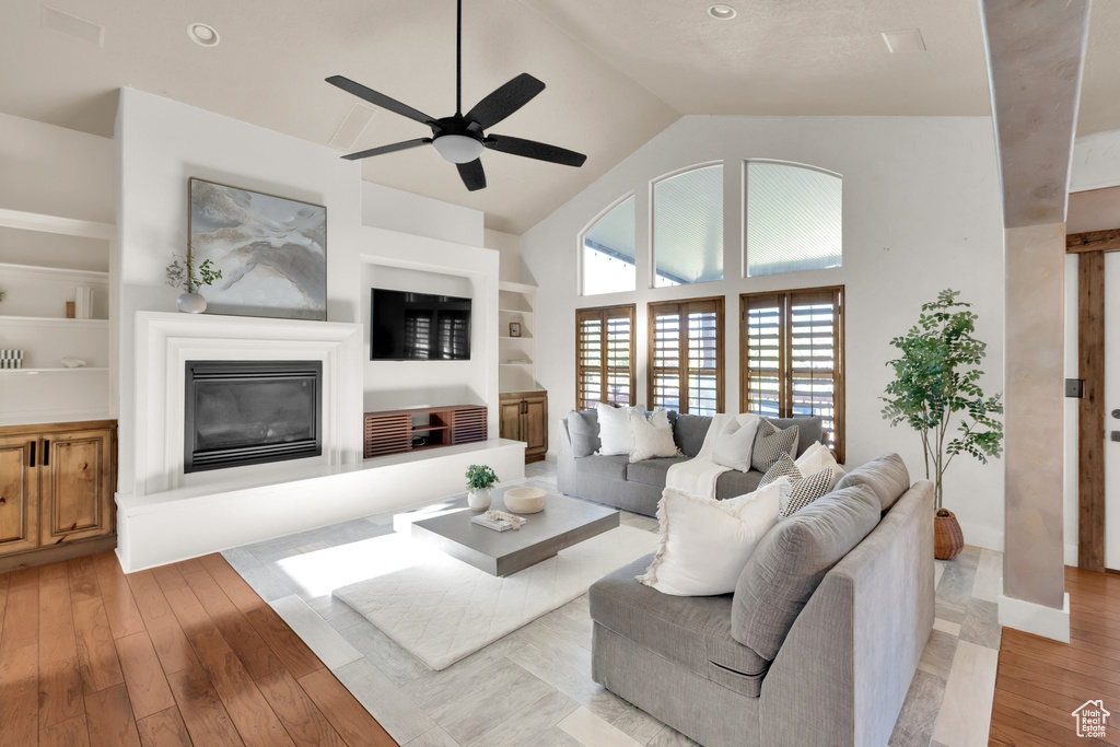Living room with a glass covered fireplace, a ceiling fan, light wood finished floors, built in shelves, and high vaulted ceiling