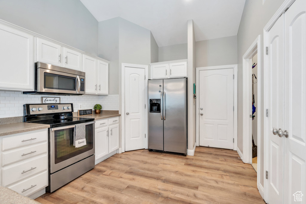 Kitchen featuring appliances with stainless steel finishes, decorative backsplash, white cabinets, light wood-type flooring, and lofted ceiling