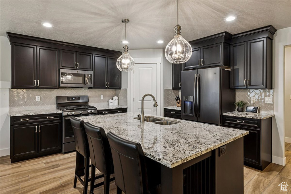 Kitchen with appliances with stainless steel finishes, a sink, light wood finished floors, light stone counters, and a center island with sink