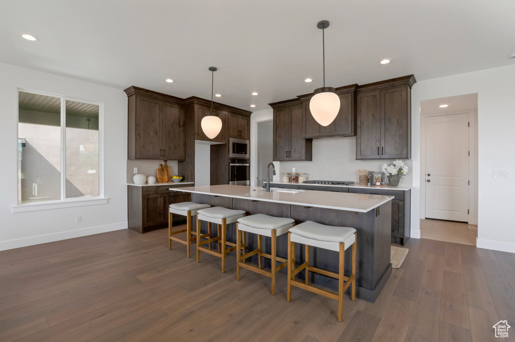 Kitchen with appliances with stainless steel finishes, dark wood finished floors, dark brown cabinetry, light countertops, and an island with sink