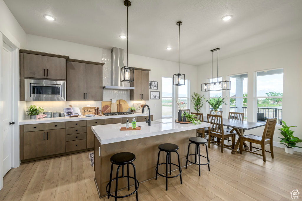 Kitchen with wall chimney range hood, stainless steel microwave, light countertops, a breakfast bar, and decorative backsplash