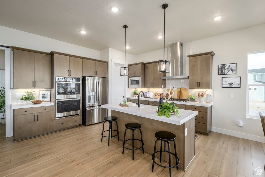Kitchen with wall chimney exhaust hood, appliances with stainless steel finishes, a breakfast bar area, light countertops, and light wood-type flooring