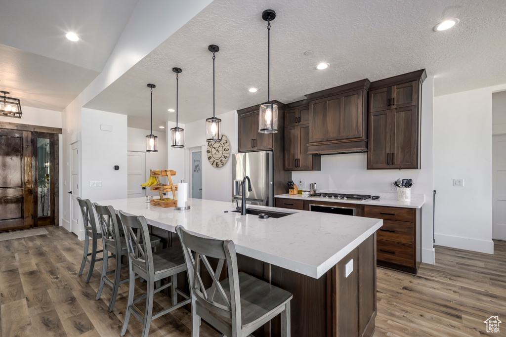 Kitchen featuring a sink, stainless steel appliances, a large island with sink, wood finished floors, and dark brown cabinets