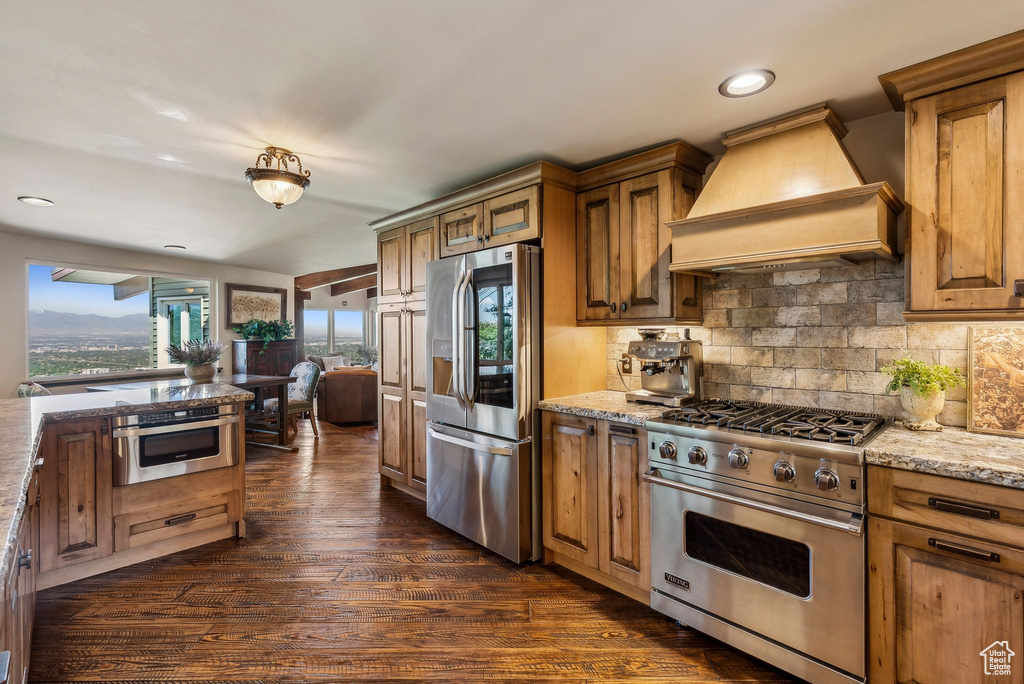 Kitchen featuring stainless steel appliances, premium range hood, dark wood-type flooring, decorative backsplash, and light stone countertops