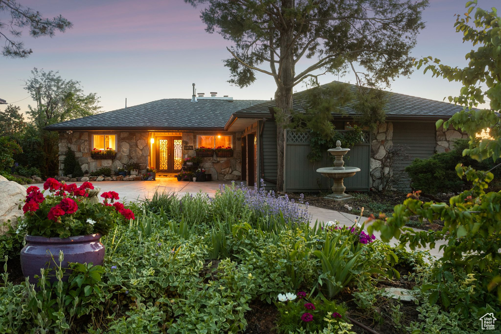 Back of house at dusk with stone siding, a patio area, and a shingled roof