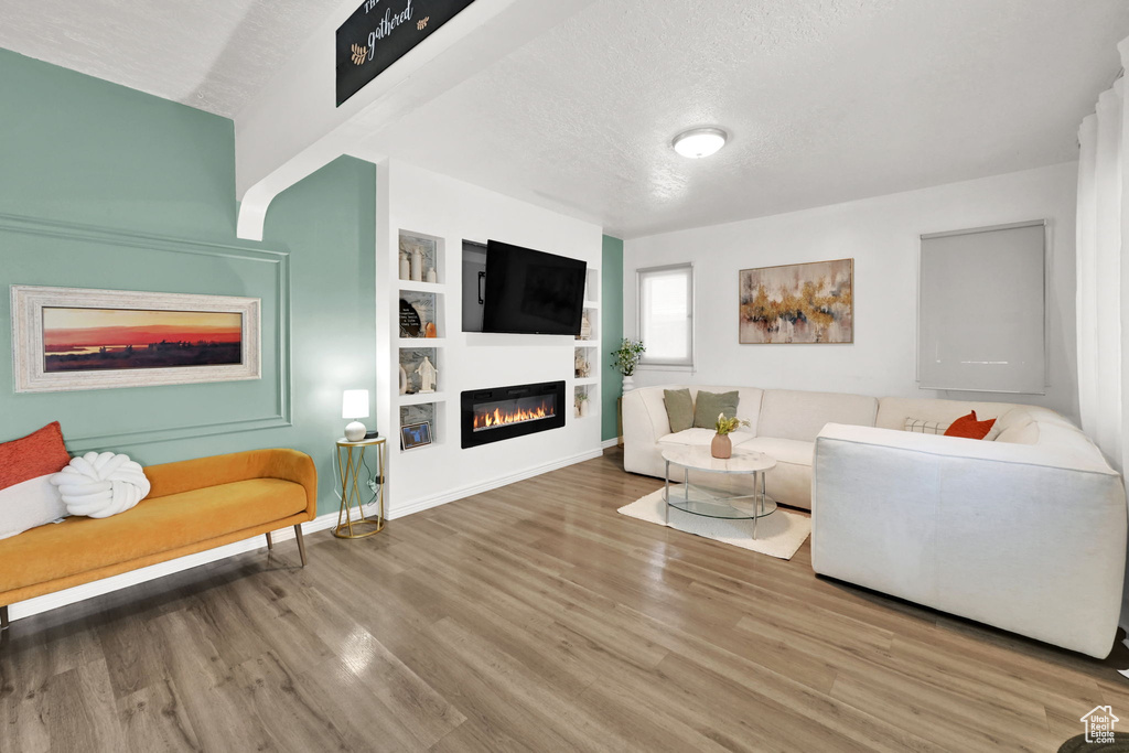 Living area featuring a glass covered fireplace, wood finished floors, a textured ceiling, built in features, and baseboards