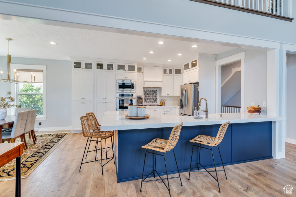 Kitchen with stainless steel appliances, a chandelier, a kitchen breakfast bar, white cabinets, and light wood-style floors