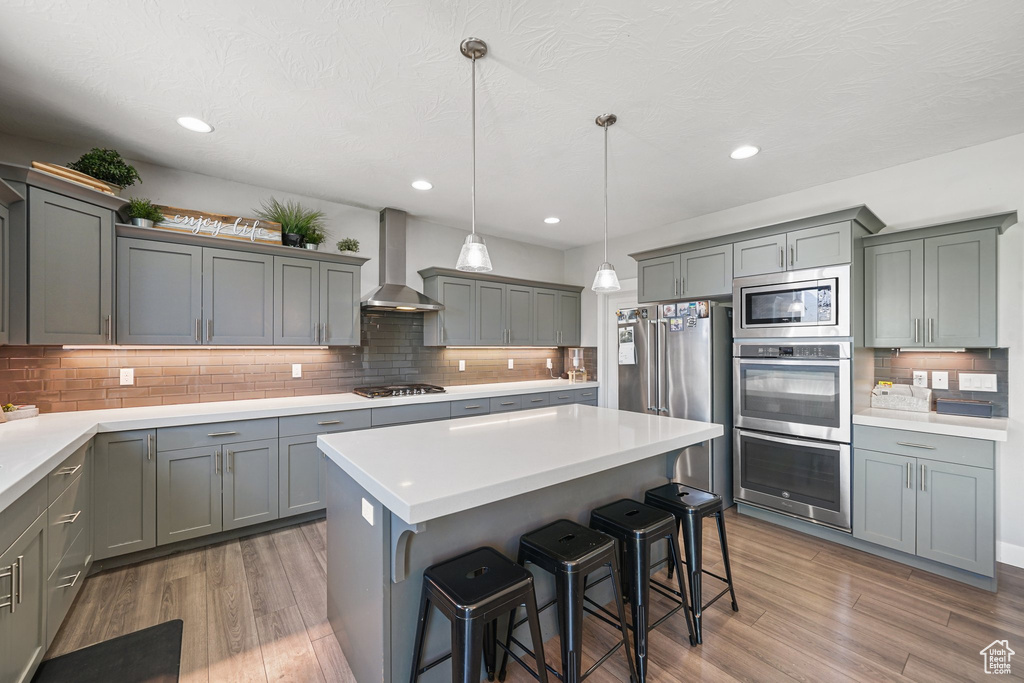 Kitchen featuring wall chimney range hood, appliances with stainless steel finishes, gray cabinetry, a kitchen bar, and recessed lighting