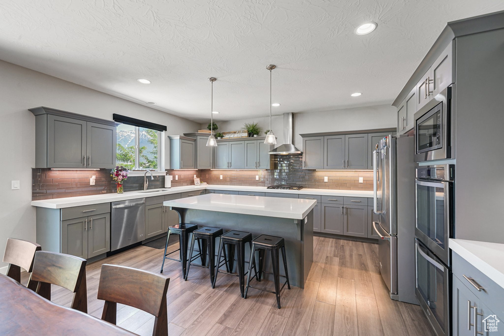 Kitchen featuring appliances with stainless steel finishes, a sink, gray cabinetry, wall chimney exhaust hood, and a kitchen island