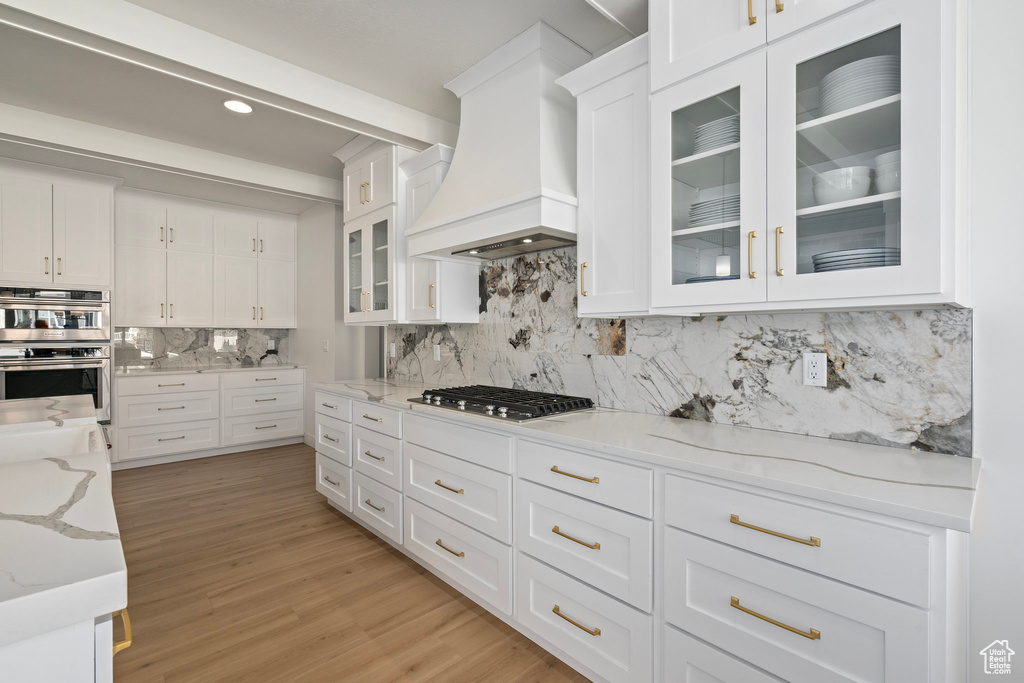 Kitchen with premium range hood, appliances with stainless steel finishes, white cabinetry, backsplash, and light stone counters