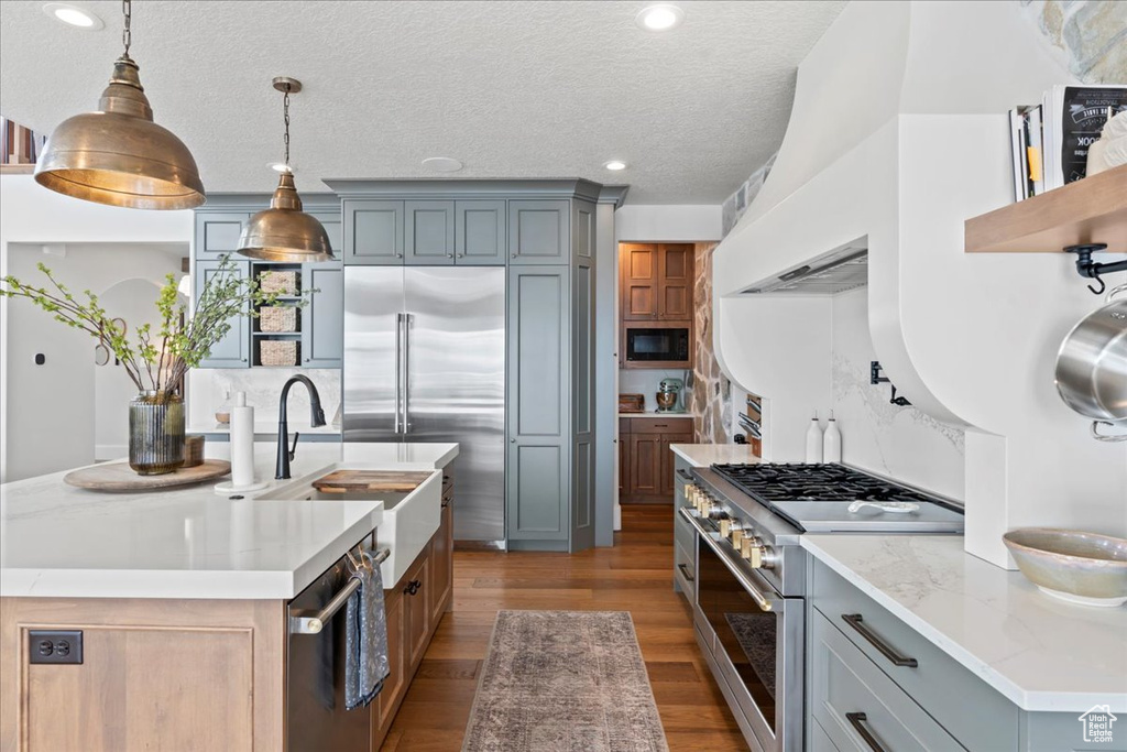Kitchen featuring built in appliances, a sink, a center island with sink, wood finished floors, and hanging light fixtures