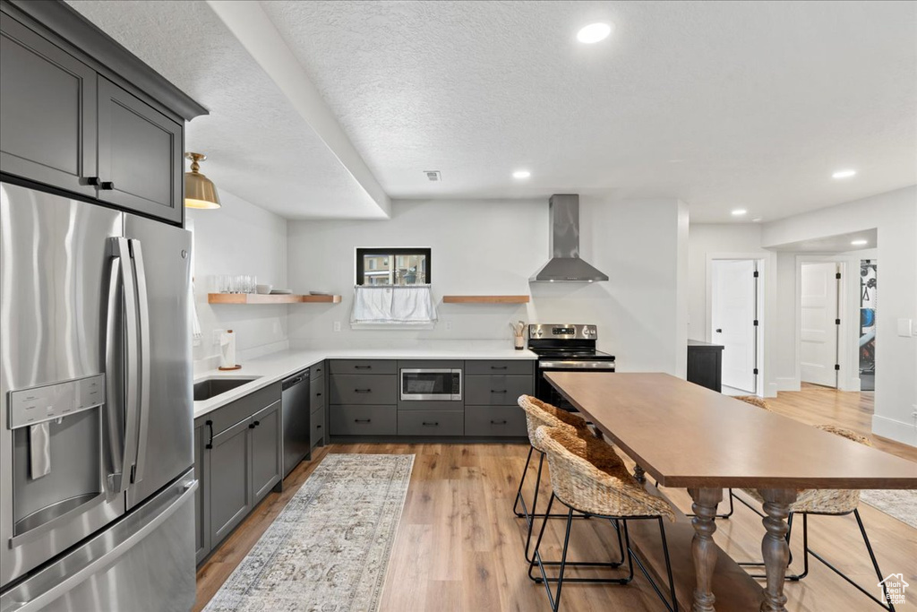 Kitchen featuring appliances with stainless steel finishes, wall chimney range hood, open shelves, gray cabinetry, and light wood-type flooring