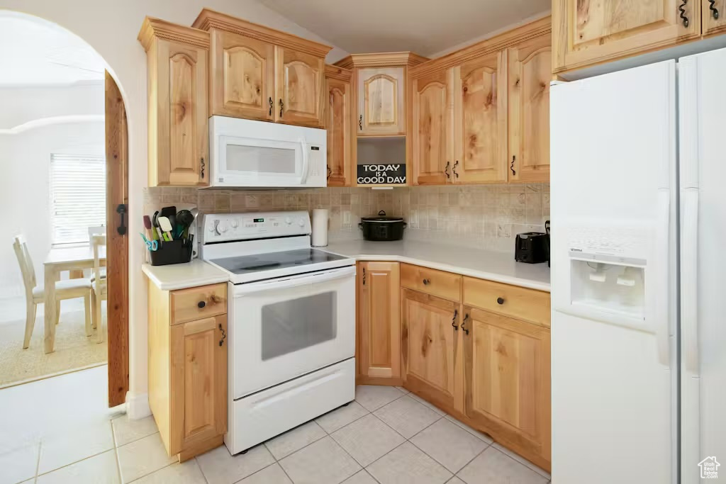 Kitchen featuring white appliances, arched walkways, light brown cabinets, tasteful backsplash, and light tile patterned flooring