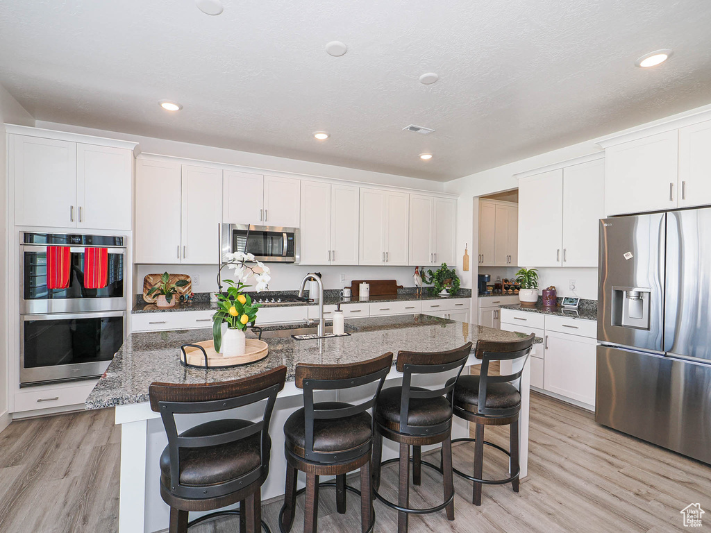Kitchen featuring appliances with stainless steel finishes, light wood finished floors, white cabinets, an island with sink, and recessed lighting
