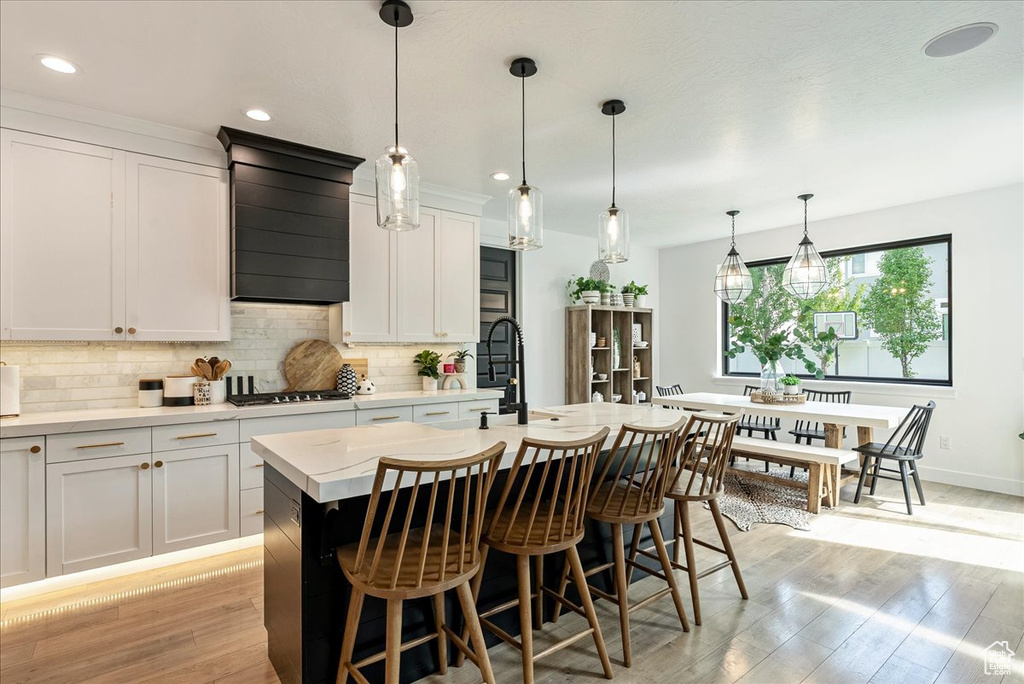 Kitchen featuring an island with sink, light wood-style floors, tasteful backsplash, a kitchen bar, and custom range hood