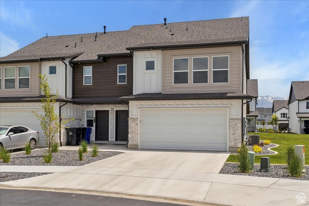 Townhome / multi-family property featuring an attached garage, driveway, stone siding, central AC, and a shingled roof